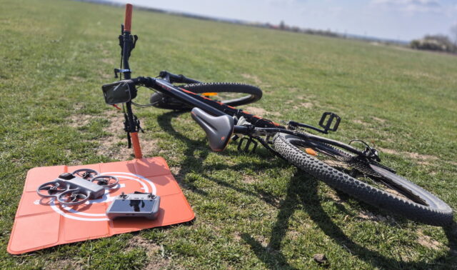 DJI Avata 360 drone flying above a trail, tracking a biker on a mountain bike in a forest setting.